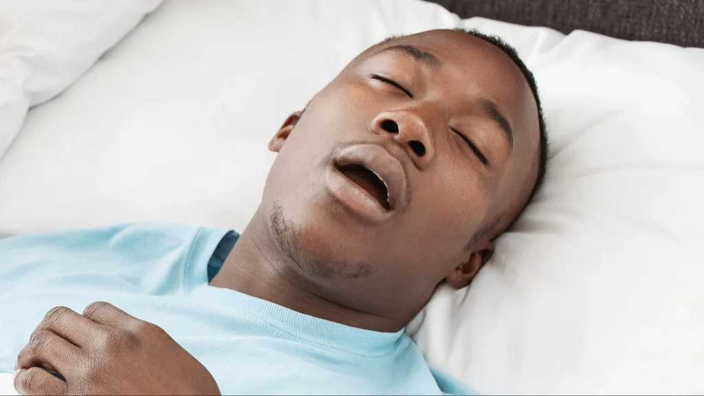 man sleeping with mouth open on a white pillow, wearing a light blue shirt, eyes closed and relaxed