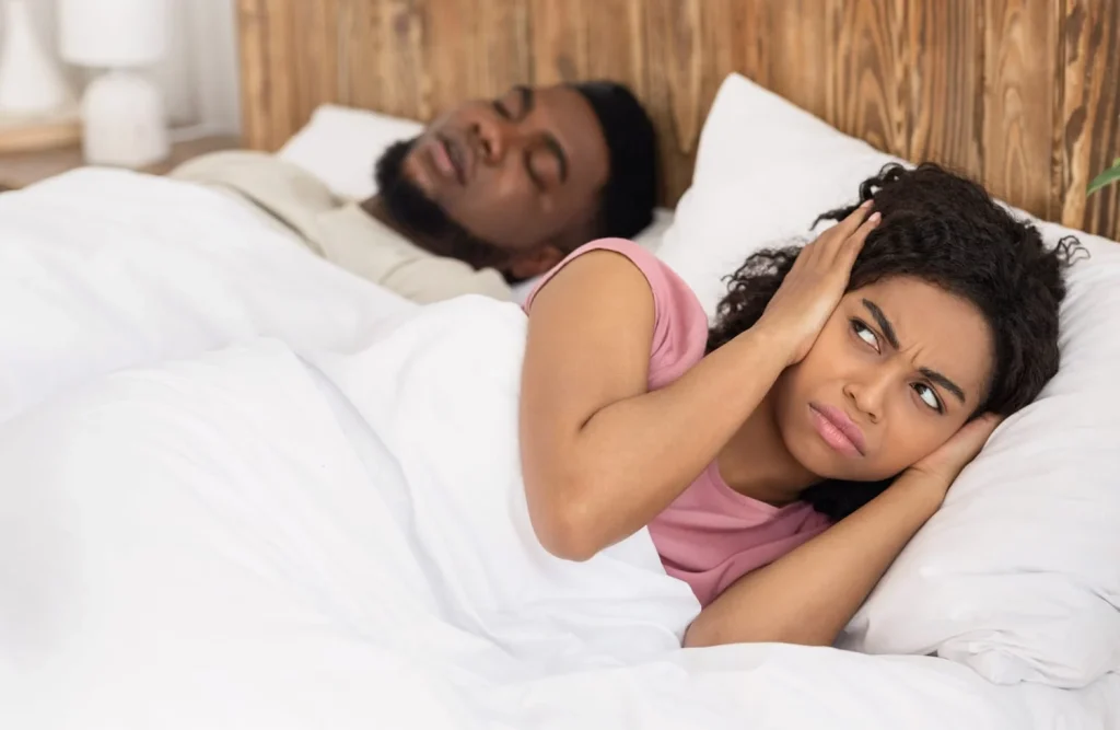 woman in bed covering her ears, looking annoyed while a man snores peacefully nearby