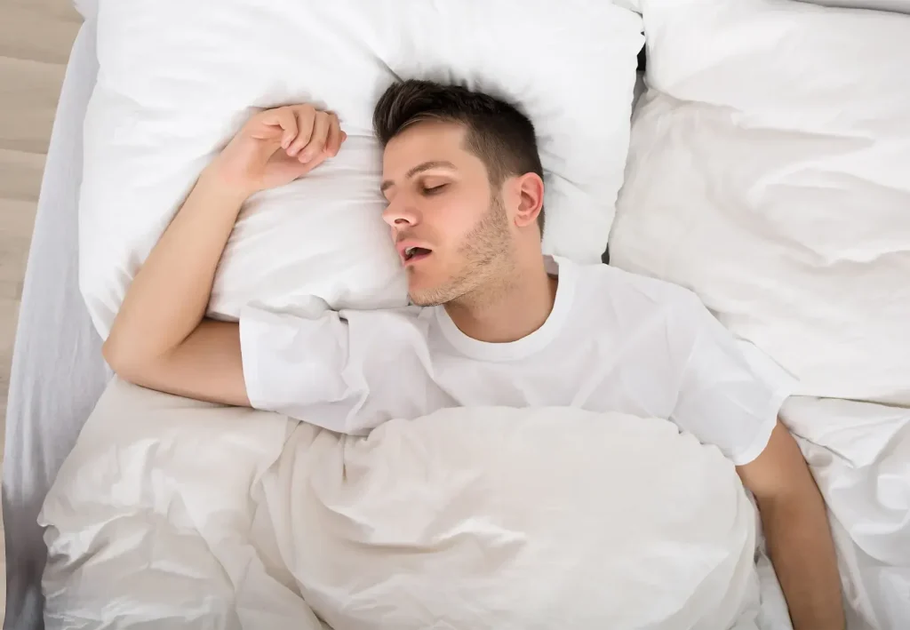 Man sleeping on a bed with white pillows, mouth slightly open, looking peaceful and relaxed.