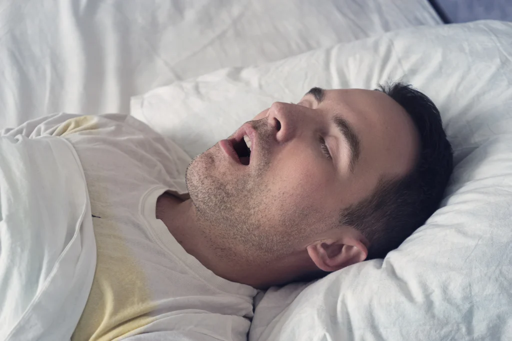 man sleeping with his mouth open on a white pillow, wearing a light-colored t-shirt