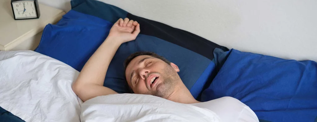 man sleeping with mouth open, surrounded by blue and white bedding, arm raised, clock on bedside table