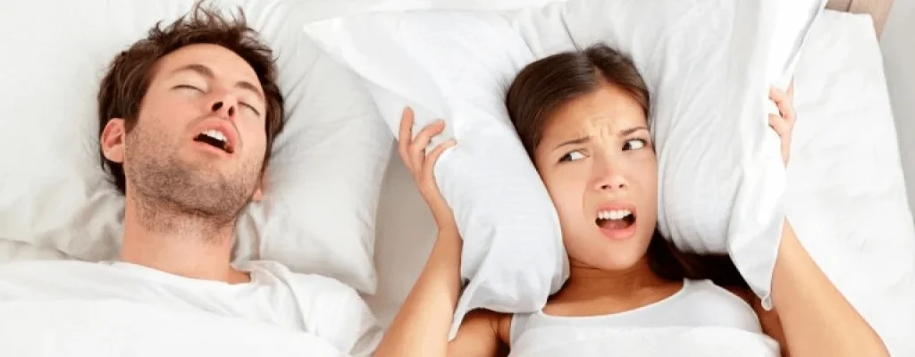 man snoring loudly while woman covers her ears, looking frustrated in bed