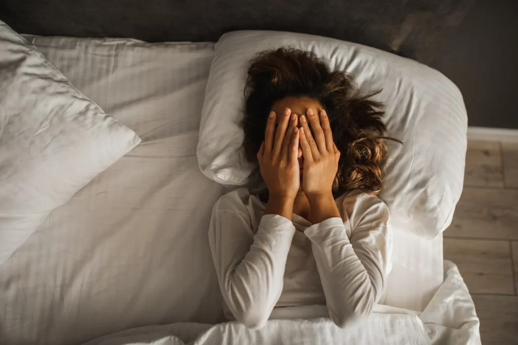 Woman lying in bed, covering her face with hands, looking distressed and unable to sleep.
