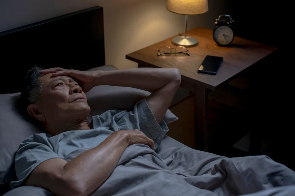 Elderly man in bed looks distressed, struggling to sleep, with a bedside lamp, clock, and glasses nearby.