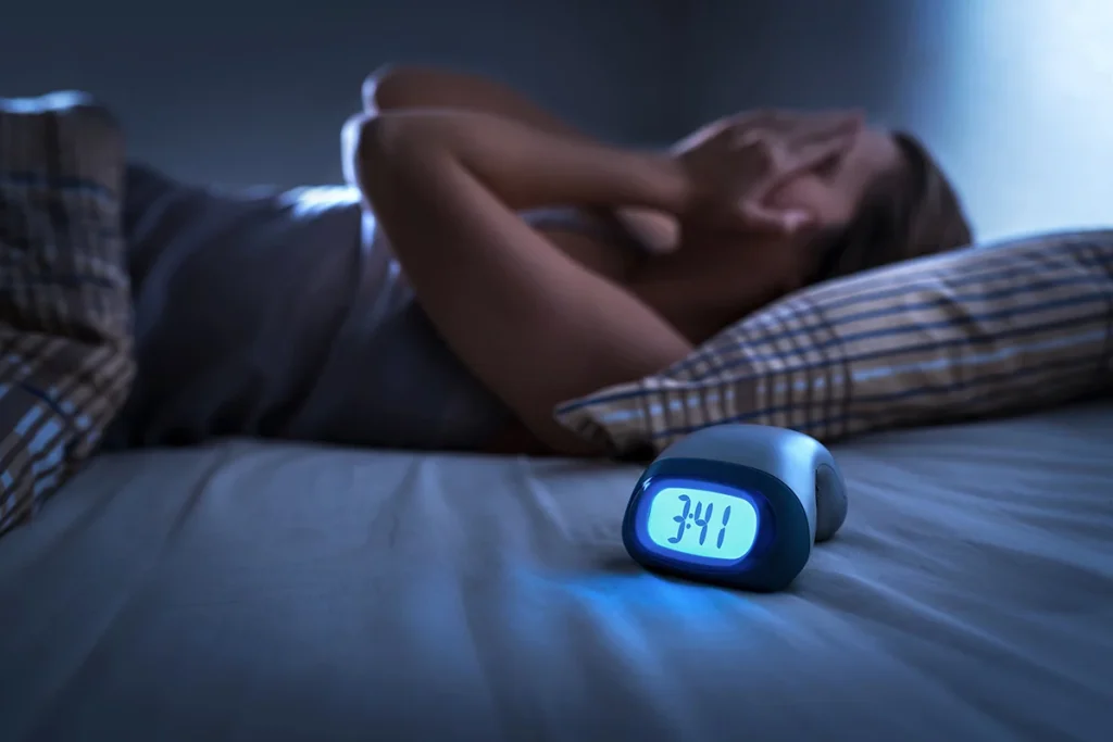 woman in bed with hands on her face, clock showing 3:41 AM in a dimly lit room