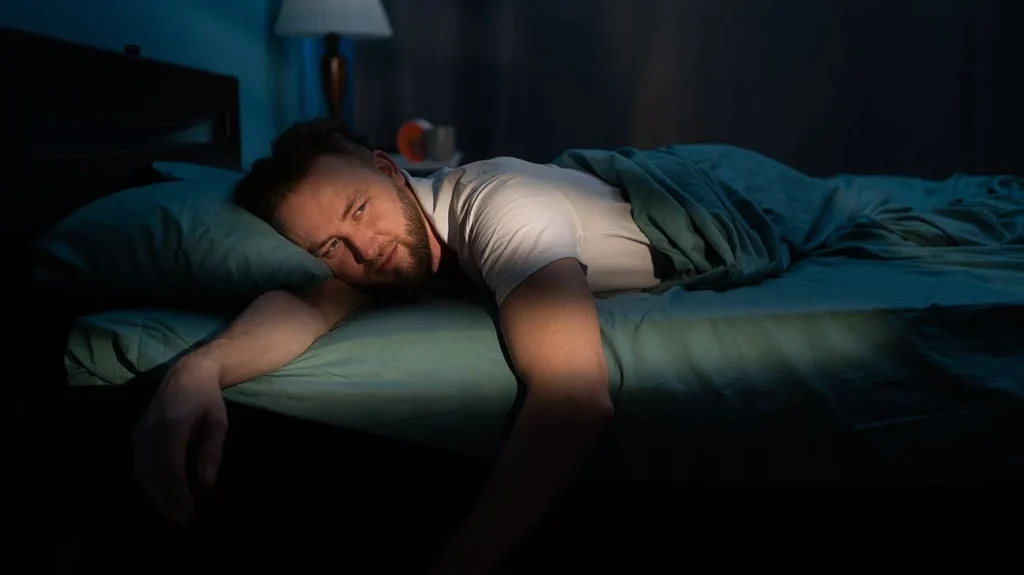 man lying in bed, looking contemplative with soft lighting and a blanket draped over him