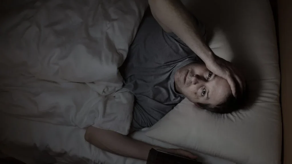 man in bed looking anxious and unable to sleep, hand on forehead, surrounded by white bedding