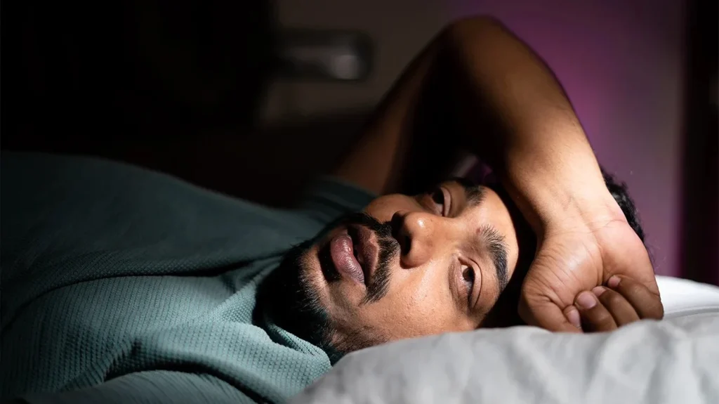 man lying in bed with a thoughtful expression, struggling to sleep in low light