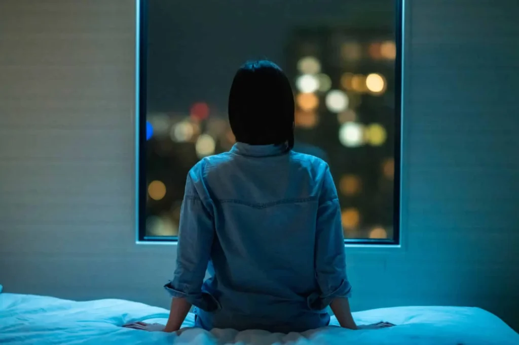 person sitting on a bed, looking out a window at a city skyline filled with colorful night lights
