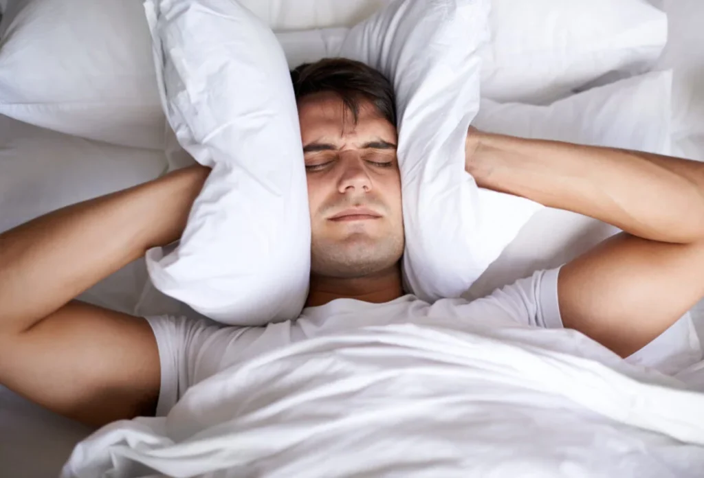 man lying in bed with pillows over his ears, appearing distressed and unable to sleep