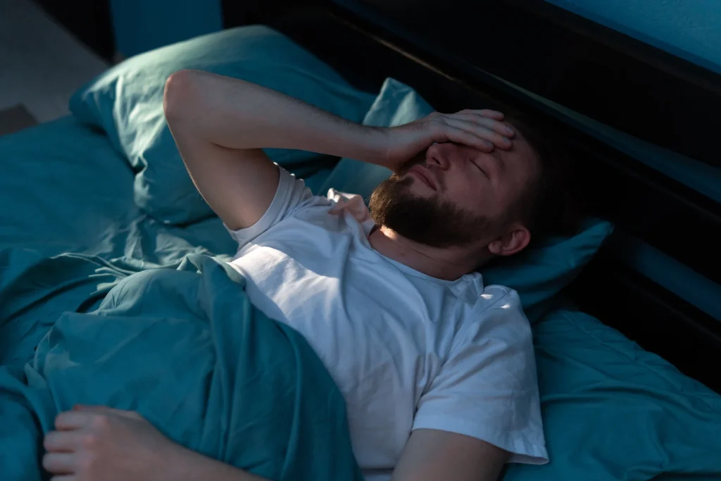 Man lying in bed, hand on forehead, looking distressed and struggling to sleep.